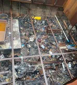 Een vervallen schuurtje waarvan het dak is ingestort, met een grote hoeveelheid afval ervoor, waaronder plastic, hout en oud gereedschap.