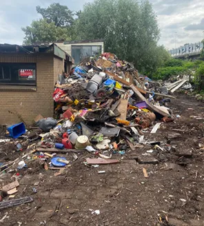 Een grote stapel schroot, oude winkelwagentjes, kabels en fietsen bij een gebouw; chaotisch opgestapeld.