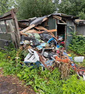 Een verroeste, verlaten boot met graffiti erop, gelegen op een overwoekerd terrein met ladders en metalen objecten.
