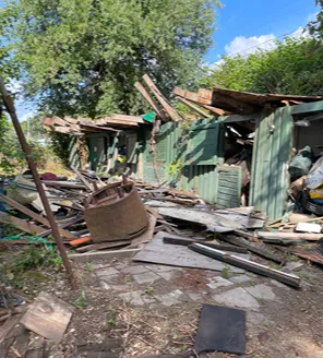 Een terrein met meerdere oude schuurtjes en verspreid afval; onder andere witgoed, hout en losse rommel liggen op de grond.