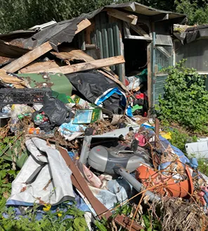 Een zwaar vervallen schuurtje met een kapot dak en rommelige omgeving vol huishoudelijk afval.