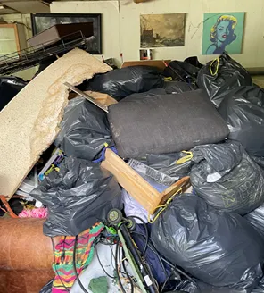 Een volledig ingestort schuurtje met puin, hout, metalen vaten en kapotte golfplaten verspreid over het terrein.