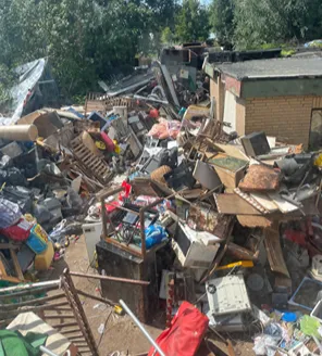 Een chaotisch terrein bezaaid met los afval, huishoudelijke apparaten, een caravan en een overdekkingszeil.