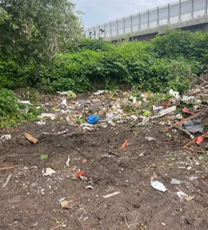 Twee grote afvalcontainers op een terrein met losliggend afval, naast een klein gebouw met een plat dak.