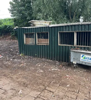 Een groen metalen gebouwtje met tralies voor de ramen. Er staat een afvalcontainer van Sita voor, en de omgeving is deels vrijgemaakt van rommel.