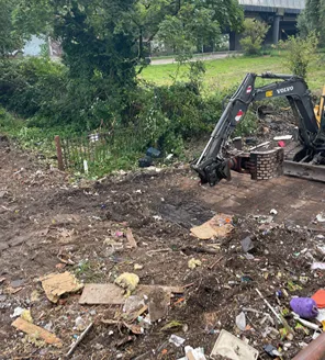 Een graafmachine is in actie op een terrein vol verspreid zwerfafval. Op de achtergrond is groen en een viaduct zichtbaar.