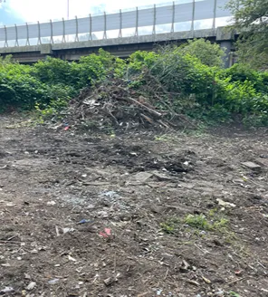 Een open ruimte met een berg snoeiafval tegen een groene achtergrond. Op de achtergrond is een viaduct met geluidsscherm zichtbaar.