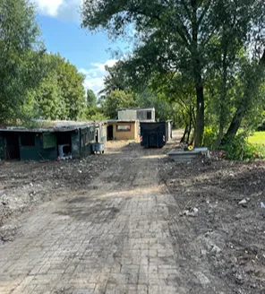 Een opnieuw betegeld pad richting enkele verouderde barakken. Het terrein is grotendeels opgeruimd, met hier en daar nog wat restafval.