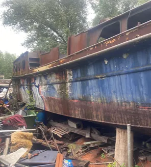 Een zwaar verroest en verlaten schip ligt tussen puin en overwoekerde begroeiing. Het vaartuig is deels gestript en omringd door wrakhout, plastic, metalen delen en afval.