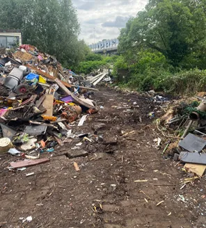 Een stuk grond achter een verwaarloosde woonunit. Er liggen afvalzakken, losse onderdelen en huishoudelijk afval, met op de achtergrond bomen.