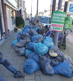 Straatbeeld met een grote berg vuilniszakken naast een verkiezingsbord.