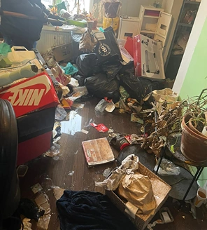 Overvolle woonkamer met vuilniszakken, kapotte planten en afval op de vloer.