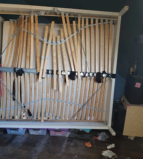 Beschadigd bedframe met gebroken latten en losse touwen, omringd door rommel en stof op de vloer.