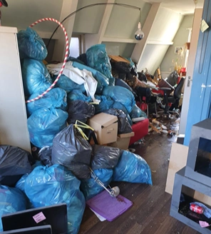 Kamer met stapels blauwe vuilniszakken, elektronica en rommel rond een bureau.