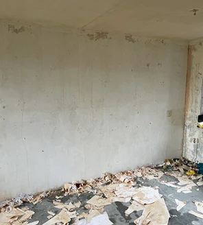 nicotine-verwijderen-en-woning-ontruimen (3) Woonkamerwand na het verwijderen van oud behang, met restanten op de vloer.