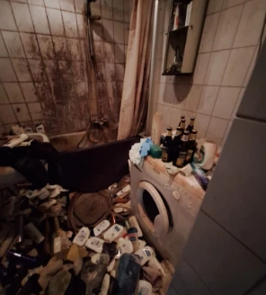 Vervuilde badkamer met wasmachine, lege flessen en stapels verzorgingsproducten op de vloer